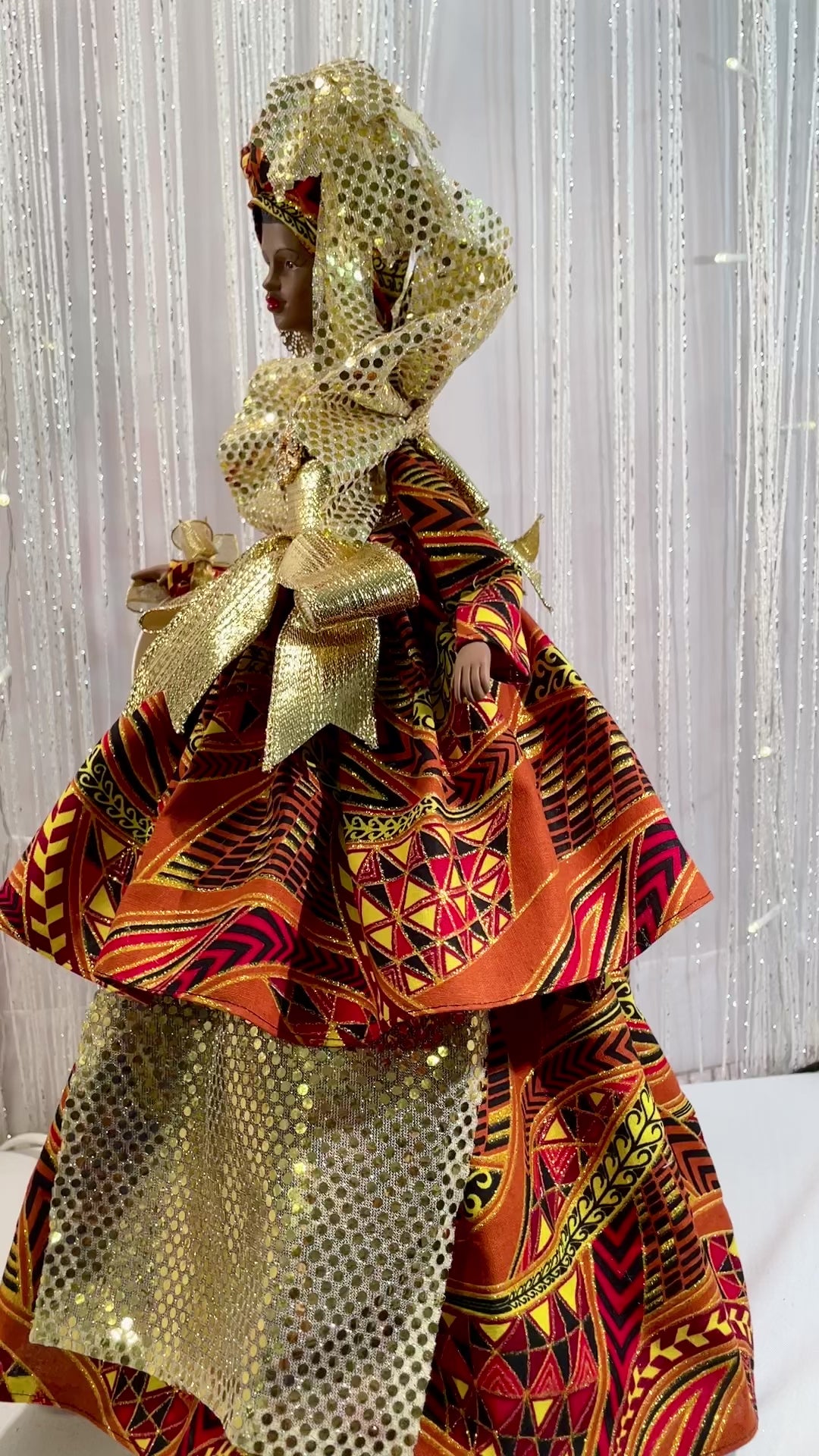 VIDEO OF AN EDUCATED BLACK QUEEN ART DOLL. She is dressed in a two tiered sparkly afrocentric fabric gown. Gold embellishments abound on this Black Queen from the collar to the waist to the draping from under her top tier. This gown is accented with a gold rhinestone cabachon at the bodice. She wears gold rhinestone earrings